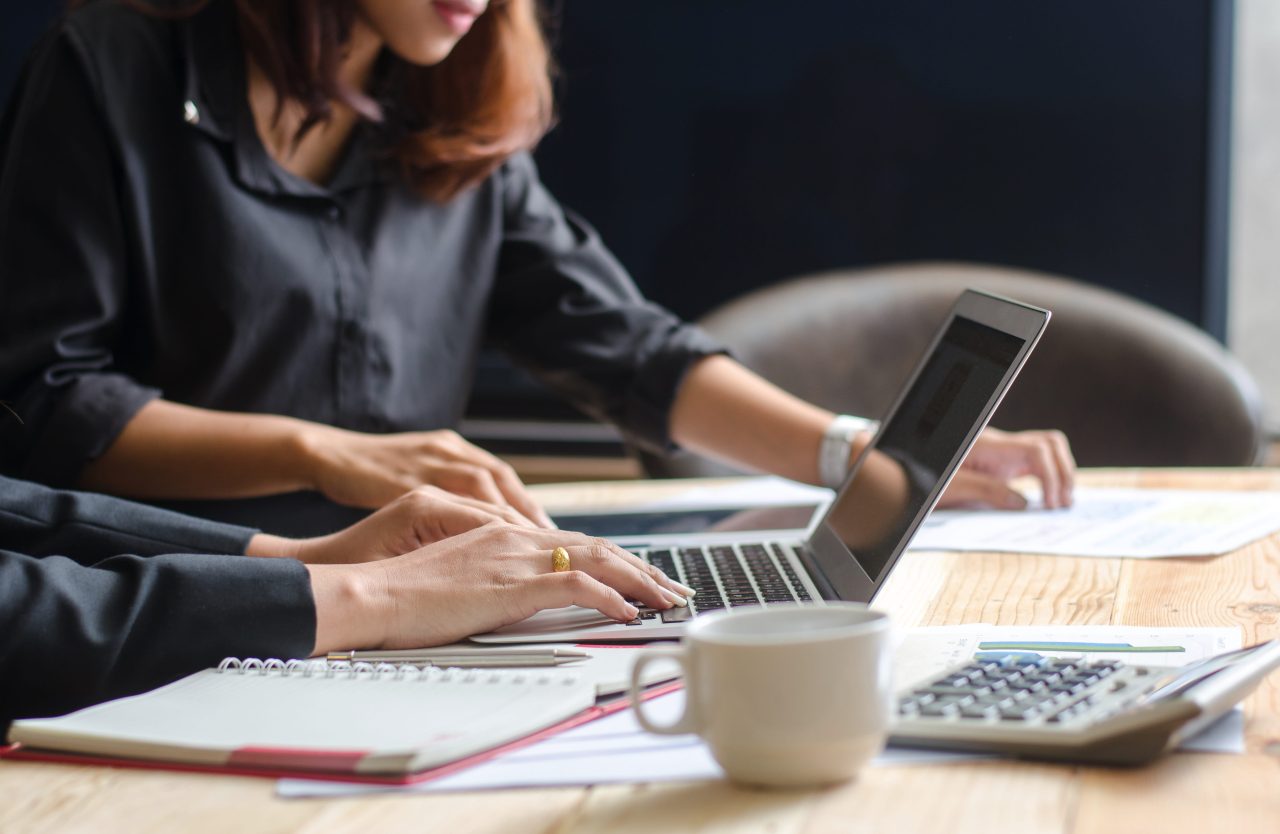 Two businesswomen collaborating on laptops at a table in a modern office setting.