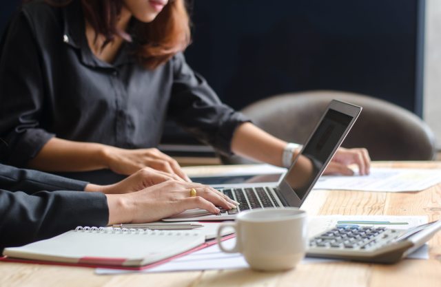 Two businesswomen collaborating on laptops at a table in a modern office setting.